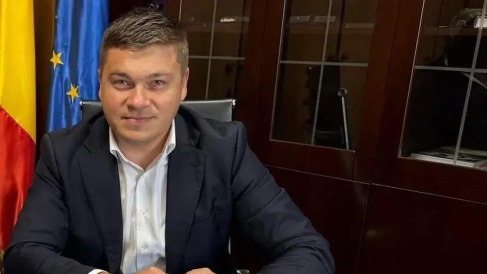 Un bărbat cu părul scurt, purtând sacou închis la culoare și cămașă albă, stă așezat în fața unui dulap de lemn și a unor steaguri.