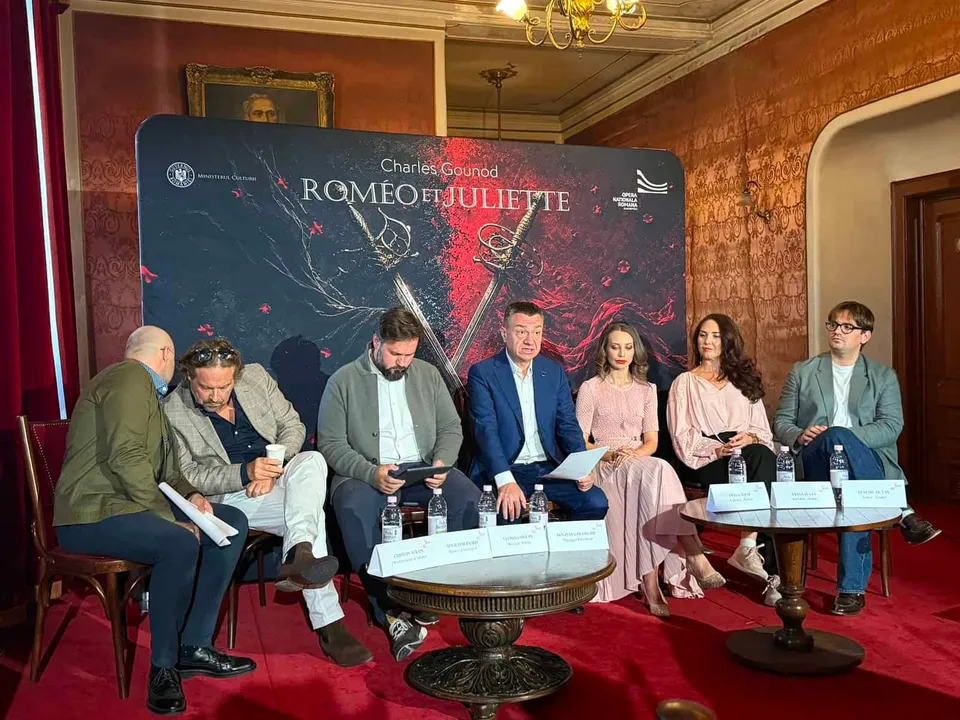 Șase persoane stau așezate la mese rotunde în fața unui panou negru pe care scrie text alb și roșu.