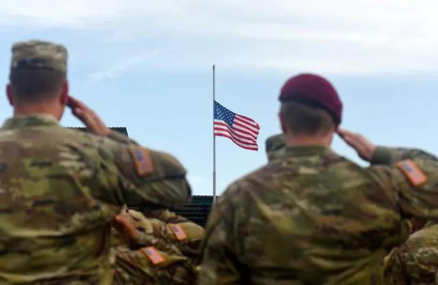 Soldați în uniforme de camuflaj stau cu spatele și execută salutul militar în fața unui steag american agățat pe un stârf sub cerul senin.