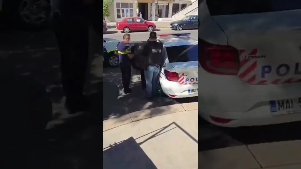 Un grup de persoane stă lângă o mașină albă cu inscripții de poliție parcată pe un trotuar lângă o clădire.