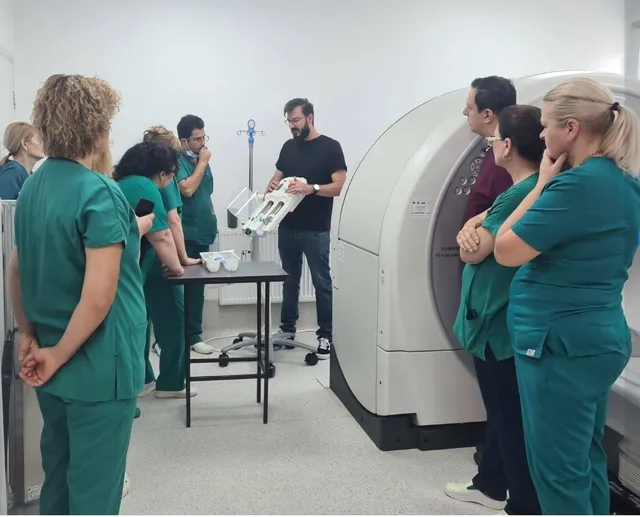 Un grup de persoane în uniforme medicale verzi privește un bărbat care ține un dispozitiv medical alb lângă o mașinărie de scanare mare.