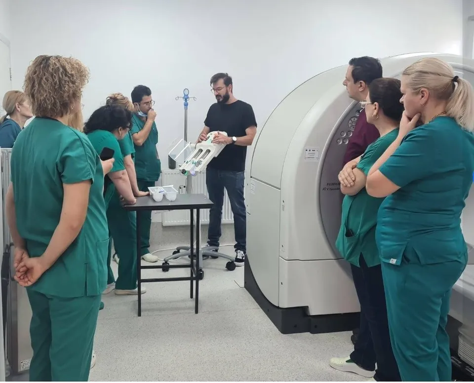 Un grup de persoane în uniforme medicale verzi privește un bărbat care ține un dispozitiv medical alb lângă o mașinărie de scanare mare.