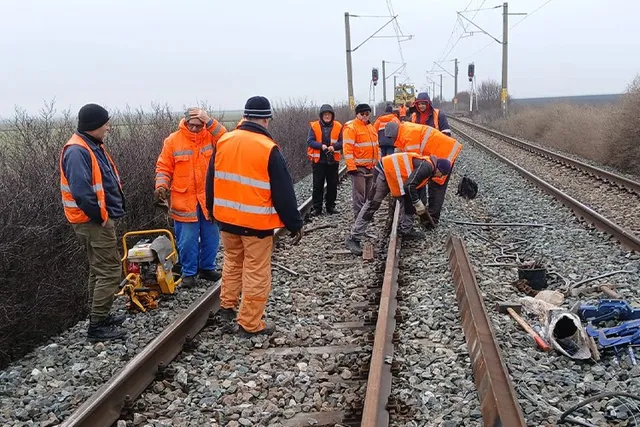 Mai mulți lucrători purtând veste de siguranță portocalie stau pe un drum feroviar lângă un echipament portabil de culoare galbenă.