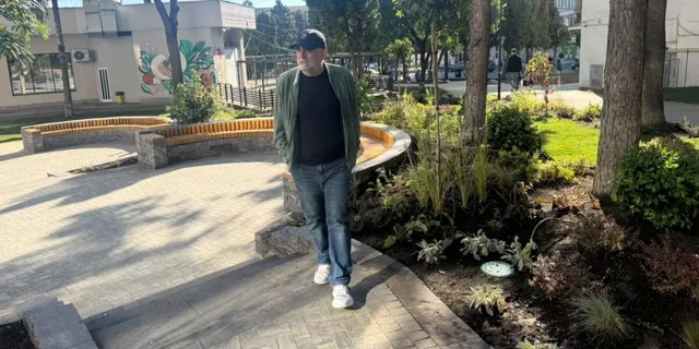 Un bărbat cu șapcă, sacou verde și bluză neagră merge pe o cărare pavată lângă un gard de piatră și plante verzi.