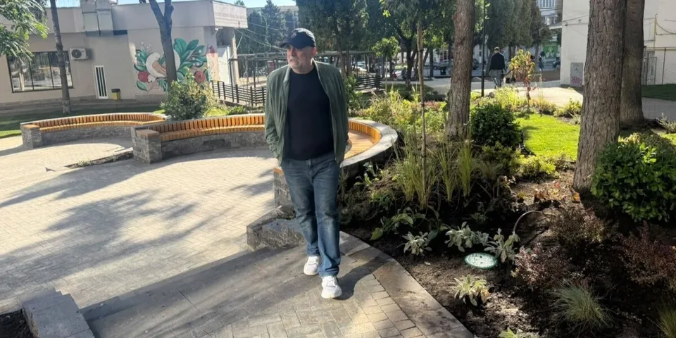 Un bărbat cu șapcă, sacou verde și blugi pășește pe o cărare pavată lângă un gard de piatră și plante verzi.