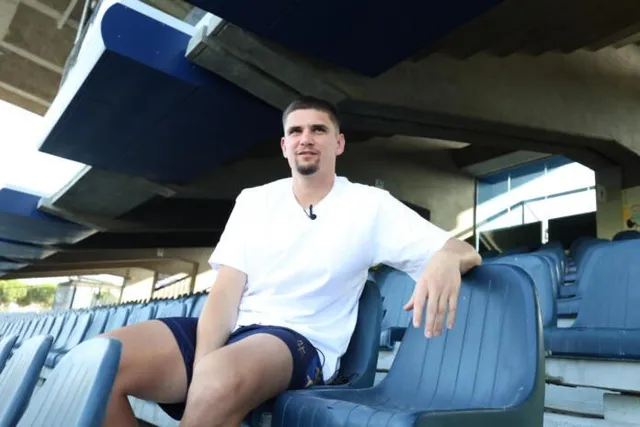 Un bărbat cu barbă scurtă și tricou alb stă așezat pe un scaun de plastic albastru într-o tribună de stadion.