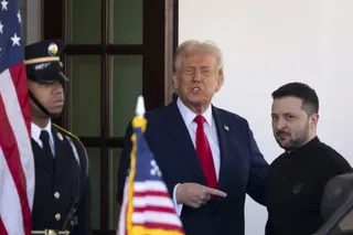 Un bărbat în costum închis cu cravată roșie și un bărbat în tricou negru stau lângă un soldat în uniformă și steaguri.