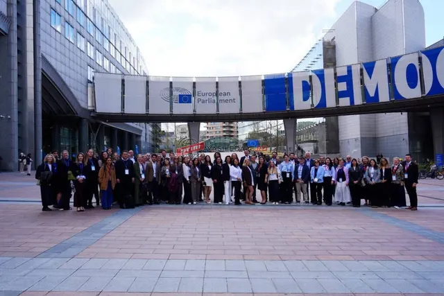 Un grup mare de oameni stă în rând în fața unui imobil modern cu un panou mare pe care scrie European Parliament și DEMOC.