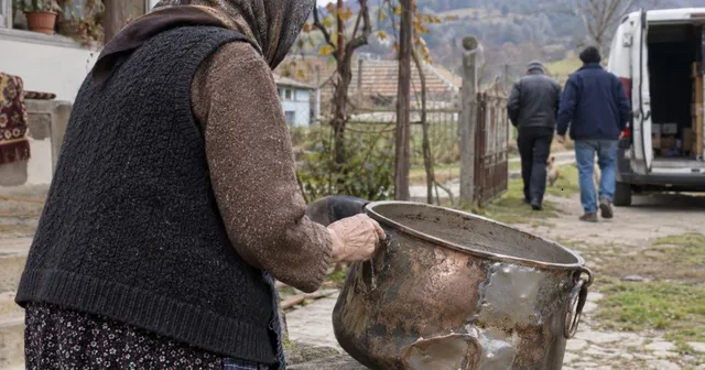 O persoană purtând un vest negru și o bluză maro ține un cazan metalic mare cu mâner negru într-un decor exterior.