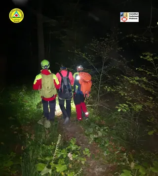 Trei persoane cu rucsacuri și echipament de munte merg pe un potecă îngustă prin pădure pe timp de noapte.