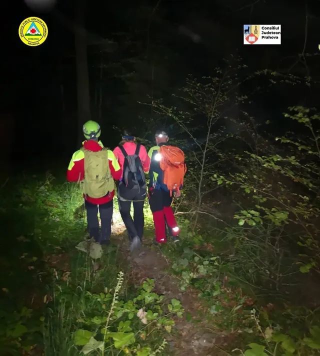 Trei persoane cu rucsacuri și echipament de munte merg pe un potecă îngustă prin pădure pe timp de noapte.