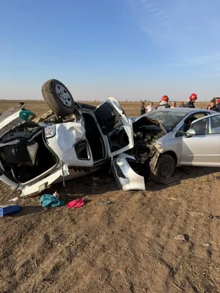 Un autoturism alb este răsturnat pe o parte pe un teren de pământ, lângă un alt vehicul gri și persoane cu casci roșii.