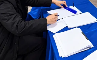 O persoană într-o jachetă neagră scrie pe o foaie de hârtie albă de pe o masă acoperită cu un material textil albastru, lângă alte foi și un obiect albastru.