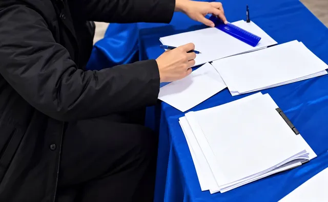 O persoană într-o jachetă neagră scrie pe o foaie de hârtie albă de pe o masă acoperită cu un material textil albastru, lângă alte foi și un obiect albastru.