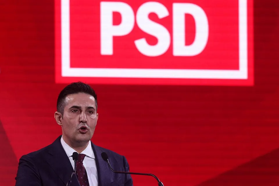 Un bărbat cu părul scurt, purtând sacou închis, cămașă albă și cravată, vorbește la microfoane în fața unui panou roșu cu literele PSD albe.