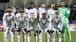 O echipă de fotbaliști îmbrăcați în echipamente albe și verzi stau grupați pe un teren de fotbal în fața unui stadion.