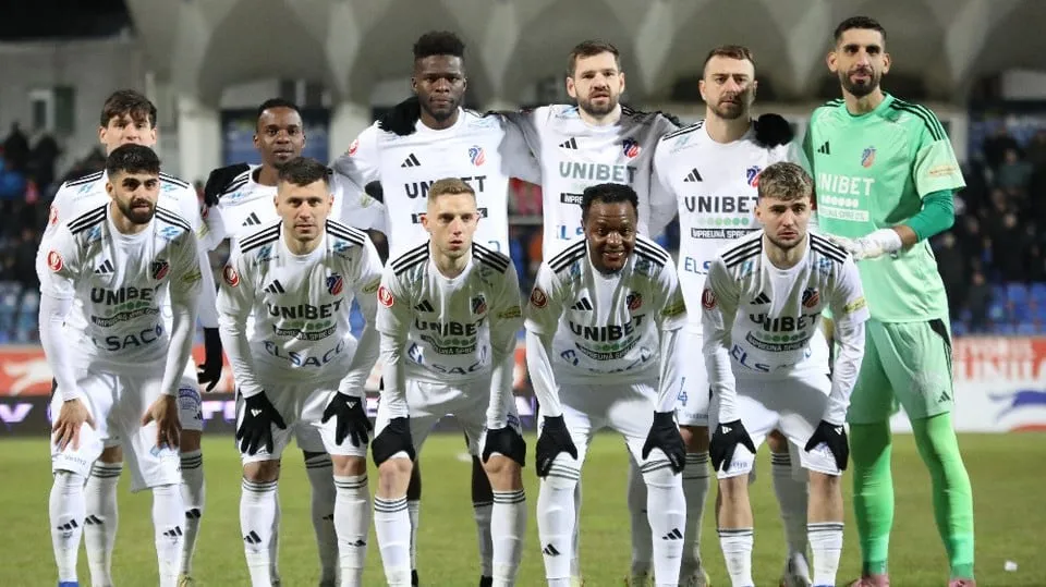 O echipă de fotbaliști îmbrăcați în echipamente albe și verzi stau grupați pe un teren de fotbal în fața unui stadion.
