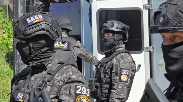 Patru persoane purtând echipament tactice negru și camuflaj, cu măști și căști protecție, stau lângă un vehicul alb cu o fereastră mică.