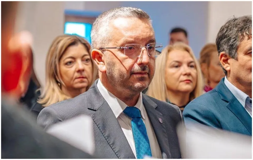 Un bărbat cu ochelari și cravată albastră stă într-o mulțime de oameni îmbrăcați în costume.
