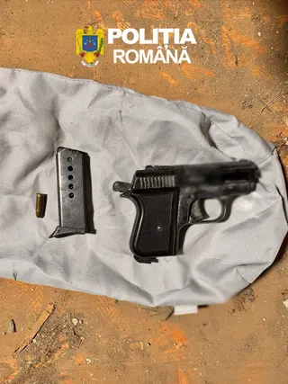 O pistolă neagră, un încărcător și un singur glonț sunt așezate pe un material textil gri deasupra unui sol nisipos.