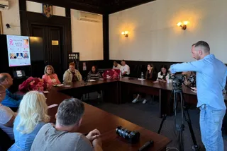 Un bărbat cu o cămașă albastră filmează un grup de persoane așezate la mese lungi într-o încăpere cu pereți albi și aplice de perete aprinse.