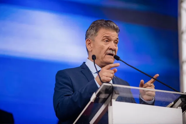 Un bărbat cu mustață, îmbrăcat în costum albastru, vorbește la un podium cu două microfoane în fața unui fundal albastru.