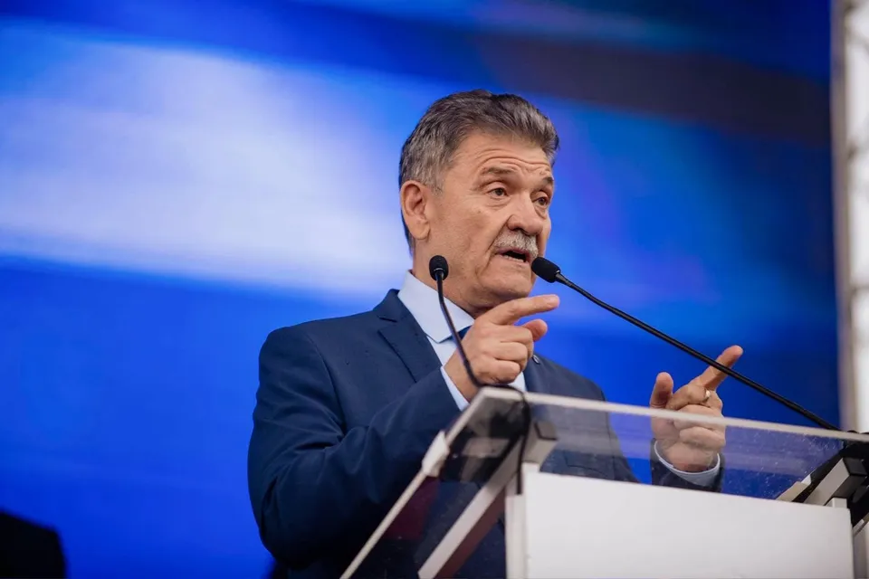 Un bărbat cu mustață, îmbrăcat în costum albastru, vorbește la un podium cu două microfoane în fața unui fundal albastru.