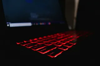 Tastatura unui laptop este iluminată de o lumină roșie într-un mediu întunecat, în timp ce ecranul afișează o interfață de sistem.