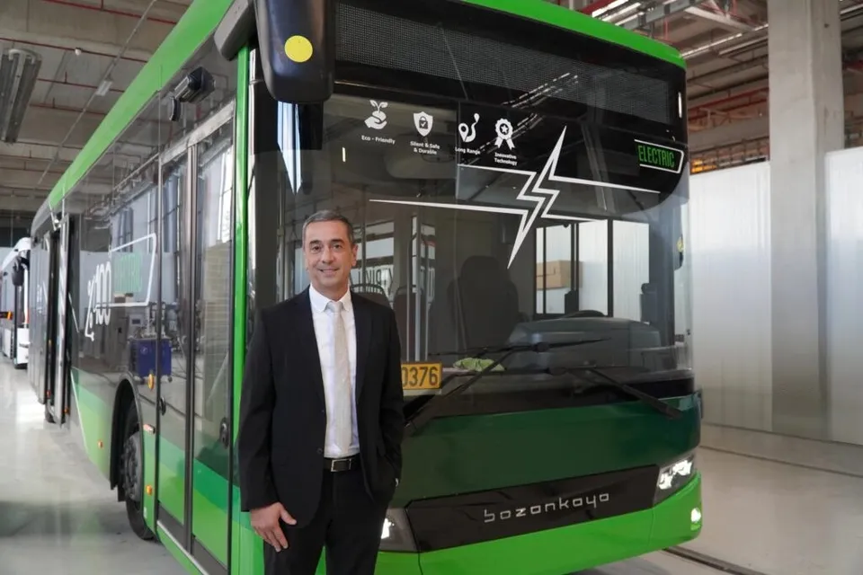 Un bărbat în costum negru și cămașă albă stă în fața unui vehicul electric verde de dimensiuni mari într-un spațiu industrial.