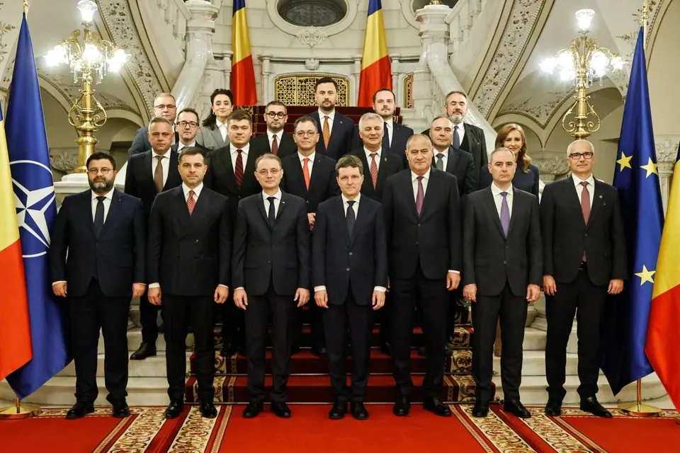 Un grup de oameni îmbrăcați în costume oficiale stă în picioare în fața unei scări interioare decorate cu steaguri și candelabre.