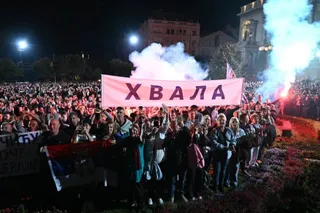 O mulțime de oameni stă în fața unui banner roz pe care scrie text în alfabet chirilic, sub un cer de noapte cu fum alb.