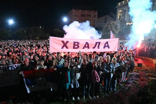 O mulțime de oameni stă în fața unui banner roz pe care scrie text în alfabet chirilic, sub un cer de noapte cu fum alb.