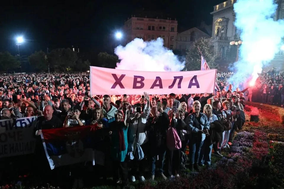 O mulțime de oameni stă în fața unui banner roz pe care scrie text în alfabet chirilic, sub un cer de noapte cu fum alb.