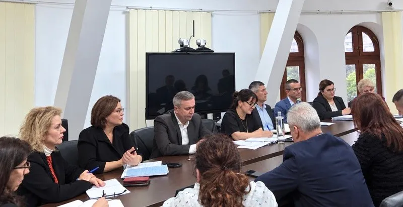 Un grup de persoane este așezat la o masă lungă într-o încăpere cu pereți albi și ferestre înalte cu rame maronii.