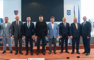 Opt bărbați în costume și haine casual stau în rând, în fața unui ecran negru și a unor blazoane pe perete.