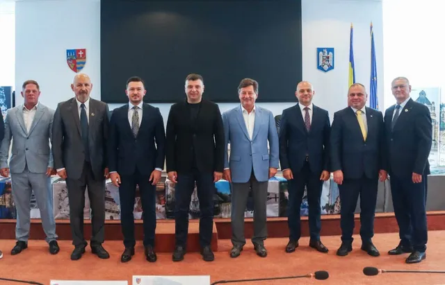 Opt bărbați în costume și haine casual stau în rând, în fața unui ecran negru și a unor blazoane pe perete.
