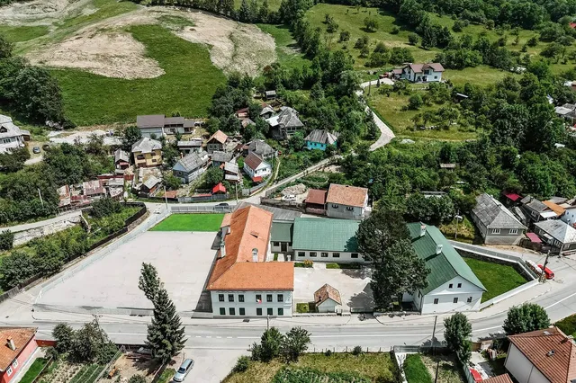 O vedere aeriană prezintă mai multe clădiri cu acoperișuri colorate, drumuri și zone verzi într-un peisaj colinar.