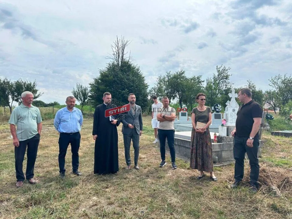 Un grup de oameni stă pe un câmp deschis lângă un monument de piatră sub un cer înnorat.