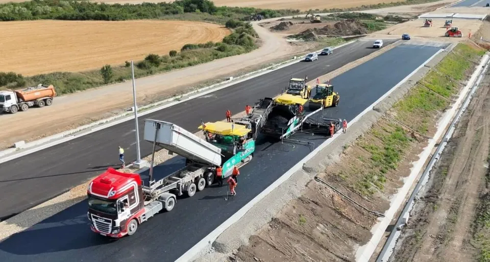 Mașini de asfalt și camioane cu remorci circulă pe o porțiune de drum proaspăt asfaltat în mijlocul unui șantier de construcții.