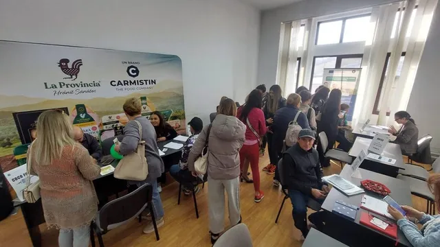 Un grup de persoane stă la mese cu computere și documente într-o încăpere cu un panou publicitar pe perete.