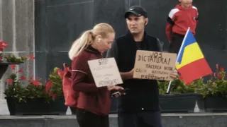 Un bărbat cu șapcă neagră și o femeie blondă țin cartoane scrise și o steag tricolor, stând în fața unui perete închis la culoare.