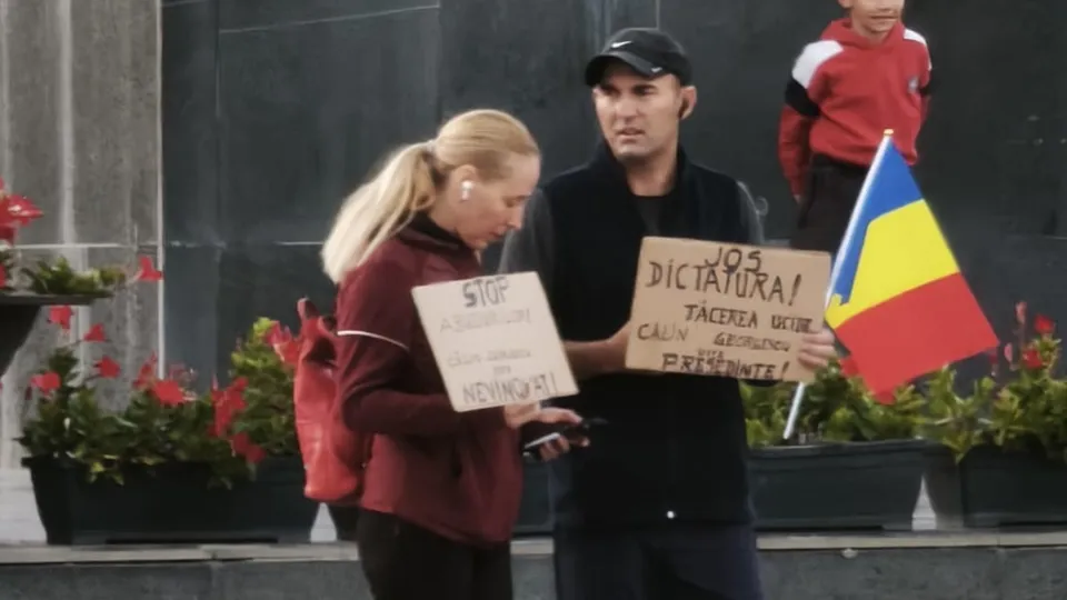 Un bărbat cu șapcă neagră și o femeie blondă țin cartoane scrise și o steag tricolor, stând în fața unui perete închis la culoare.