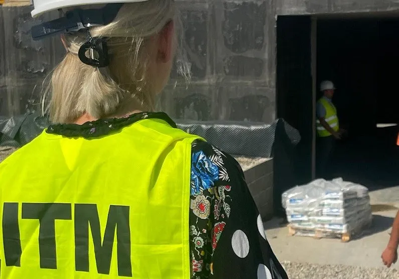 O persoană cu părul blond poartă o vestă reflectorizantă galbenă cu inscripția ITM și privește spre o intrare de clădire cu un alt om îndepărtat.