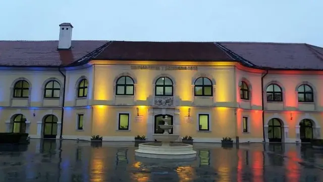 O clădire galbenă cu acoperiș înclinat este iluminată cu lumini colorate pe o suprafață umedă sub un cer închis la culoare.