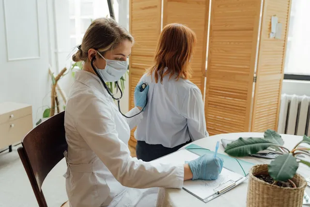 O femeie cu halat alb și mască medicală folosește un stetoscop, în timp ce o altă femeie stă cu spatele la cameră lângă o masă rotundă.
