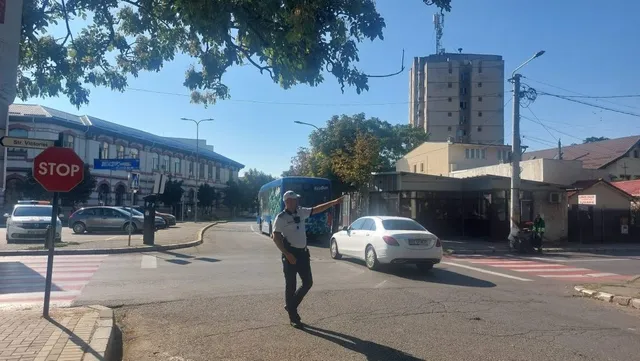 Un polițist cu uniformă deschisă stă pe drum și indică spre lateral, lângă un vehicul alb și un autobuz albastru sub cerul senin.