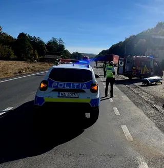O mașină de poliție albastră și albă stă pe marginea unui drum asfaltat lângă un camion de pompieri și un om cu vestă reflectorizantă.