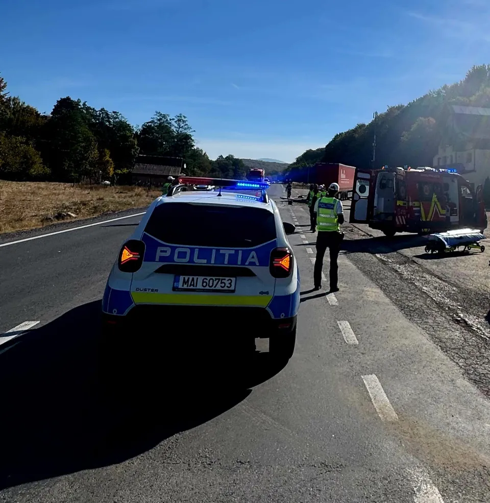 Un vehicul de poliție albastru cu girofare aprinse stă pe marginea unui drum asfaltat lângă un camion de pompieri și un operator vestit în verde.