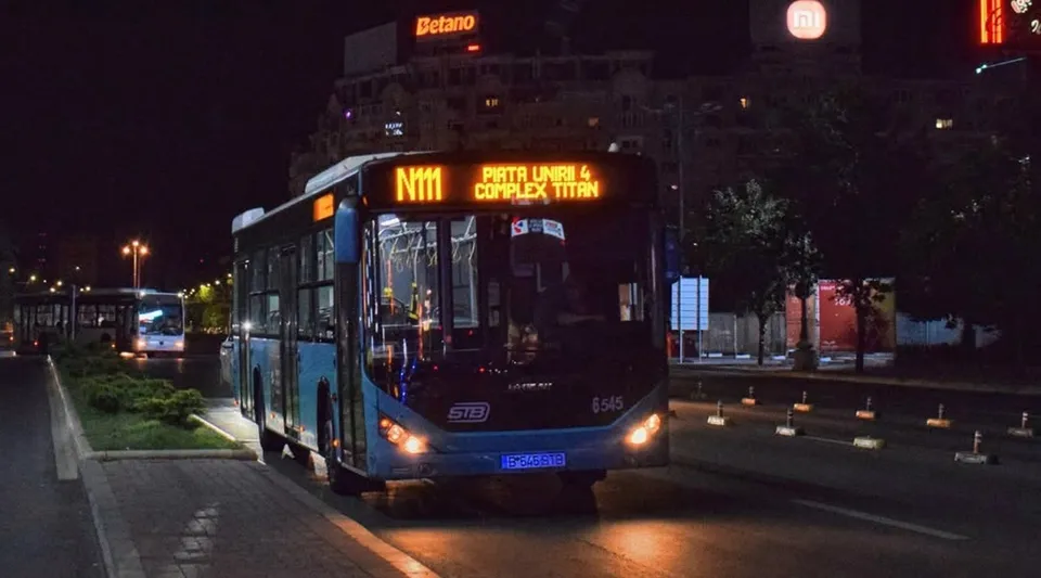 Un autobuz albastru cu afișaj digital N111 circulă pe un drum asfaltat noaptea, având alte vehicule și clădiri iluminate în fundal.