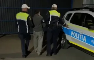 Doi polițiști în uniformă cu benzi reflectorizante conduc un bărbat într-o direcție lângă un autoturism de poliție albastru și alb.